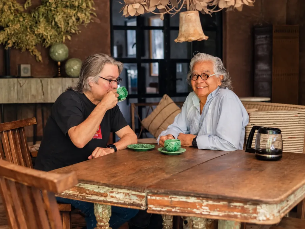 Couple sharing coffee at a weathered wooden table in the dining room, beneath a wrought-iron chandelier.